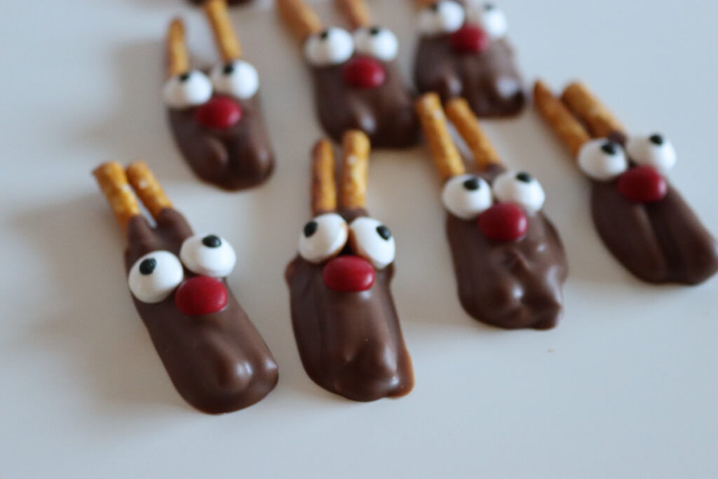 Chocolate-dipped cookies decorated with candy eyes, red candy noses, and pretzel sticks, arranged to resemble reindeer on a white surface.