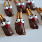 Chocolate-dipped cookies decorated with candy eyes, red candy noses, and pretzel sticks, arranged to resemble reindeer on a white surface.