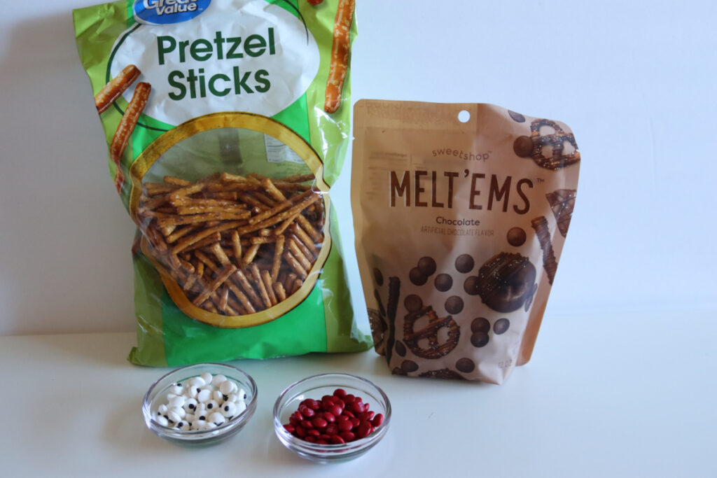 A bag of pretzel sticks, a bag of chocolate melt candies, a bowl of candy eyeballs, and a bowl of red candies displayed on a white surface.