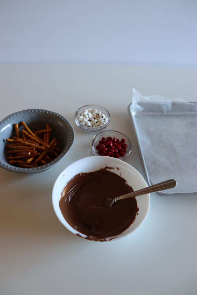 A bowl of melted chocolate with a spoon, pretzel sticks, red candies, white candy eyes, and a baking tray lined with parchment paper on a white surface.