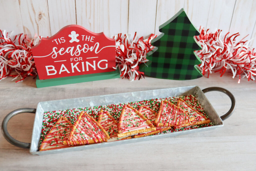 Rectangular tray with Christmas tree-shaped cookies decorated with red and green icing and sprinkles, in front of holiday-themed decor and a sign reading "'Tis the season for baking.