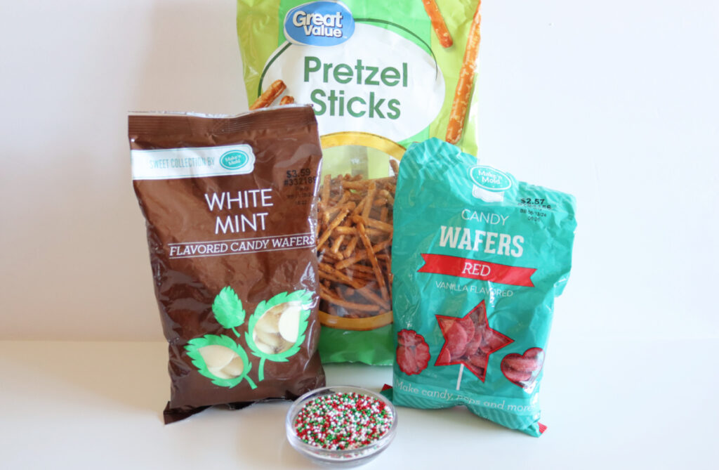 A bag of pretzel sticks, a bag of white mint candy wafers, a bag of red candy wafers, and a small dish of round holiday sprinkles on a white surface.