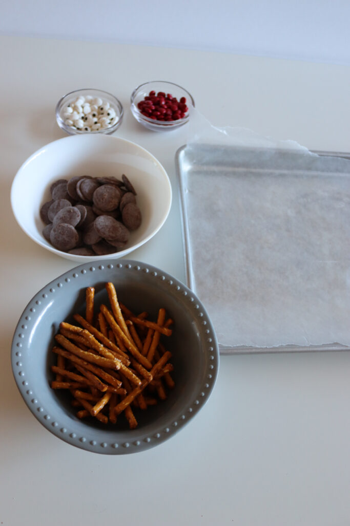 A baking sheet lined with parchment, a bowl of pretzel sticks, chocolate wafers, and small bowls of red and white candies are arranged on a white surface.
