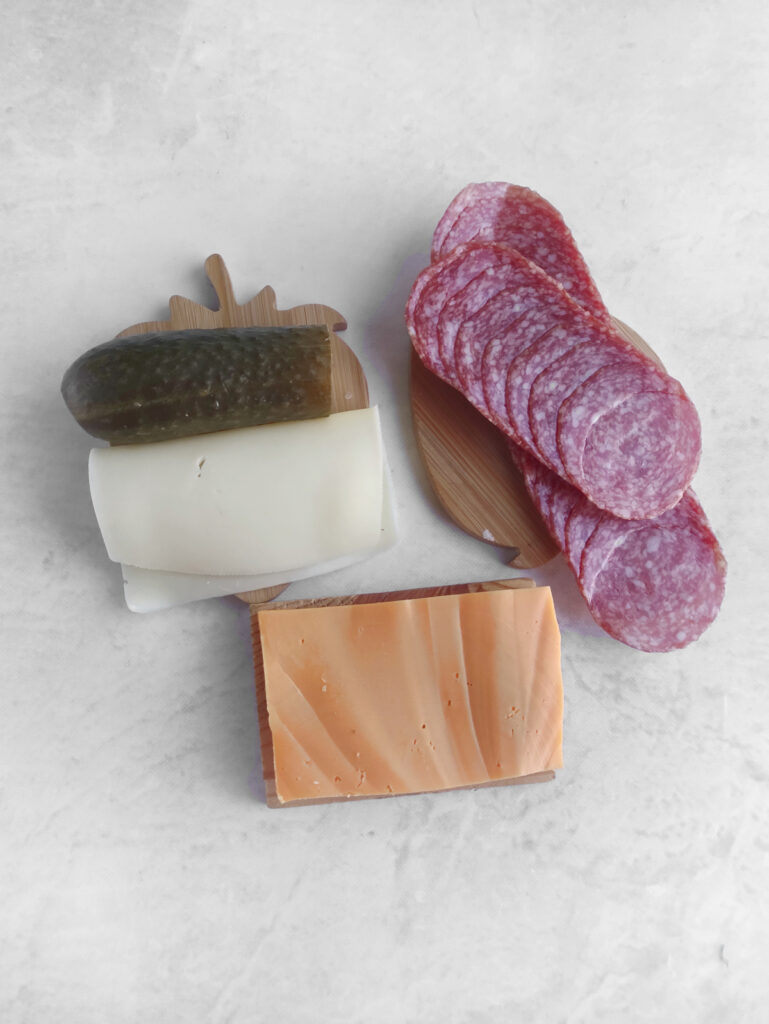 Slices of salami, a stack of white cheese with a pickle, and a block of orange cheese are arranged on a light-colored surface.