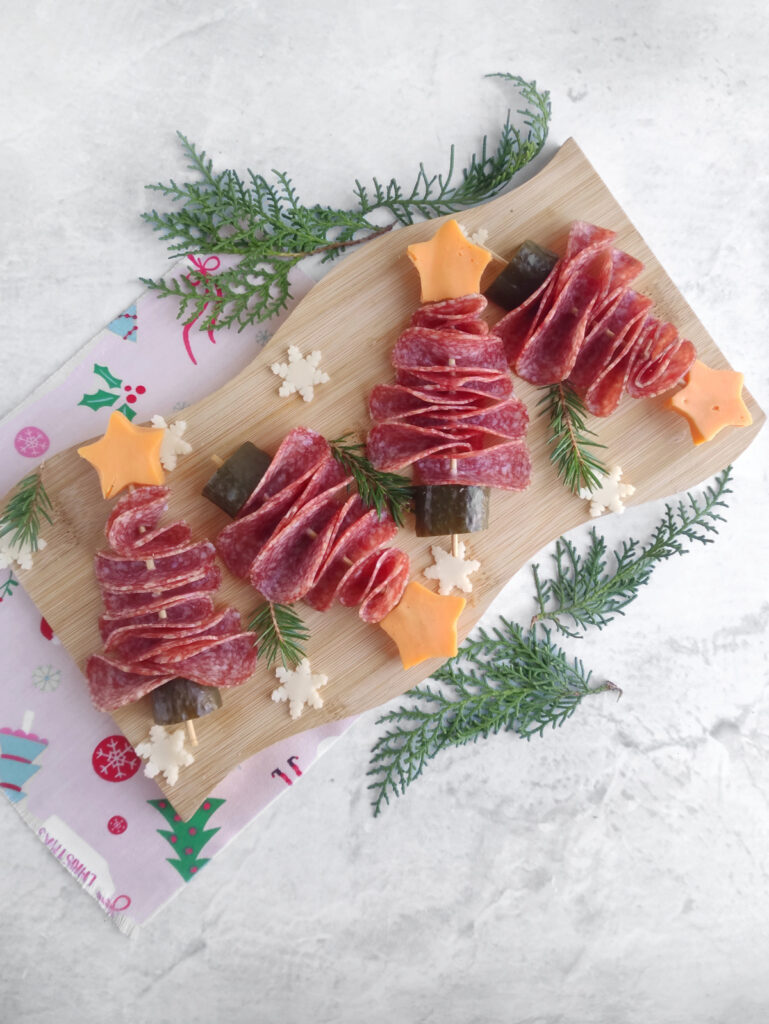 A wooden board with salami arranged as Christmas trees, decorated with cheese stars and snowflakes, and garnished with herbs on a holiday-themed napkin.