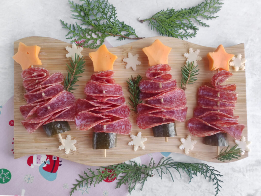 Four Christmas tree-shaped appetizers made of folded salami, cucumber bases, and cheese stars on skewers, arranged on a wooden board with greenery and small cheese snowflakes.