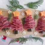 Four Christmas tree-shaped appetizers made of folded salami, cucumber bases, and cheese stars on skewers, arranged on a wooden board with greenery and small cheese snowflakes.