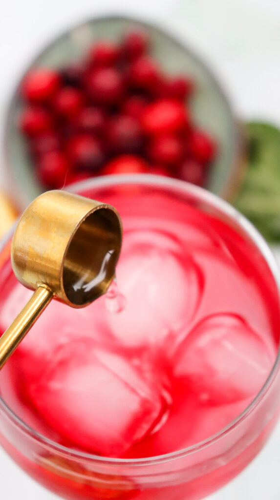A close-up of a gold jigger pouring liquid into a glass of pink drink with ice, with a bowl of cranberries in the background.