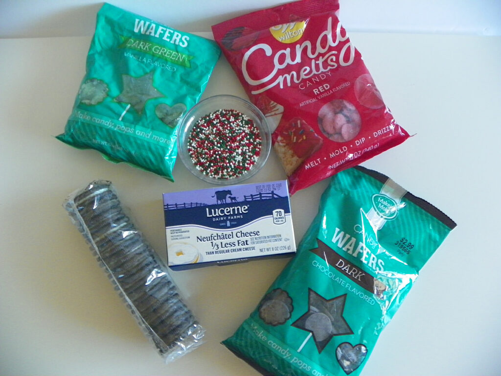 An assortment of baking ingredients: chocolate wafers, candy melts, sprinkles, a package of cream cheese, and a package of sandwich cookies on a white surface.