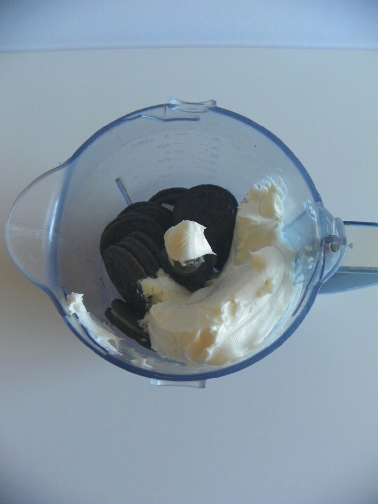 A blender cup containing whole chocolate sandwich cookies and a large amount of cream cheese, ready for mixing.