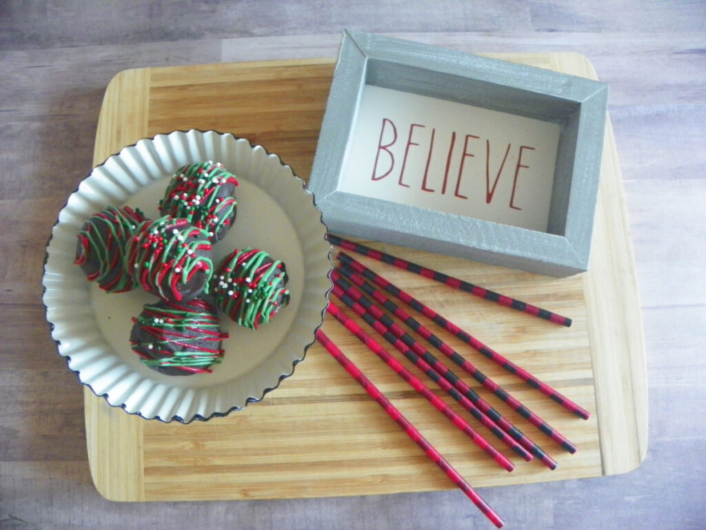 A plate with four chocolate-covered treats and a framed sign reading "BELIEVE" sit on a wooden board, alongside several red and black striped straws.