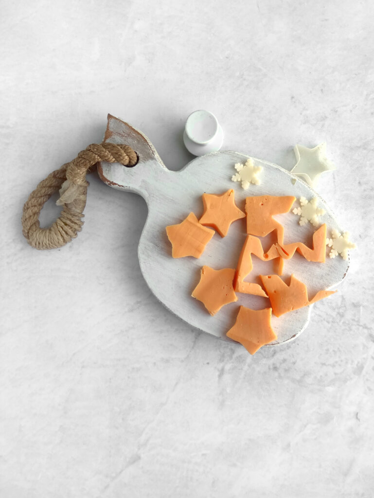 A white cutting board with a rope handle holds star-shaped cheese and white flower-shaped cheese pieces on a light gray surface.