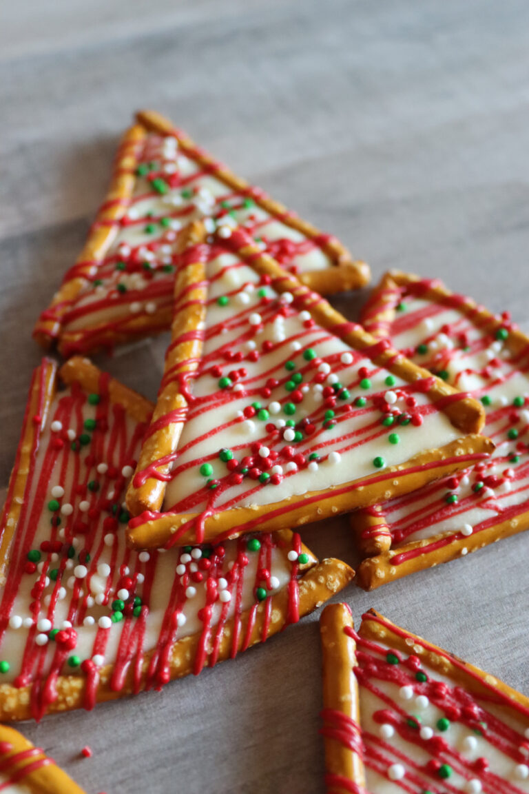 Triangle-shaped holiday cookies decorated with white icing, red drizzle, and red, green, and white sprinkles, bordered by pretzel sticks, arranged on a wooden surface.
