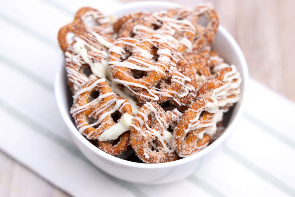A white bowl filled with pretzels coated in cinnamon sugar and drizzled with white icing sits on a striped cloth.
