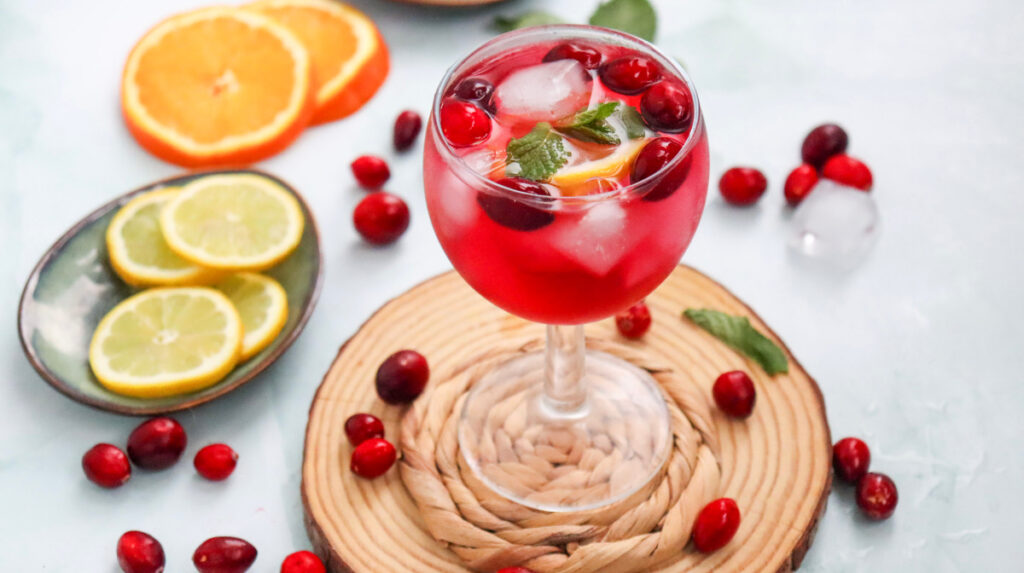 A glass of red fruit punch with ice, mint, cranberries, and lemon slices, surrounded by cranberries, orange slices, and lemon slices on a light surface.