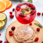 A glass of red fruit punch with ice, mint, cranberries, and lemon slices, surrounded by cranberries, orange slices, and lemon slices on a light surface.