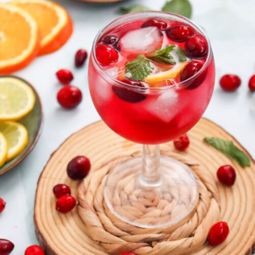 A glass of red fruit punch with ice, mint, cranberries, and lemon slices, surrounded by cranberries, orange slices, and lemon slices on a light surface.