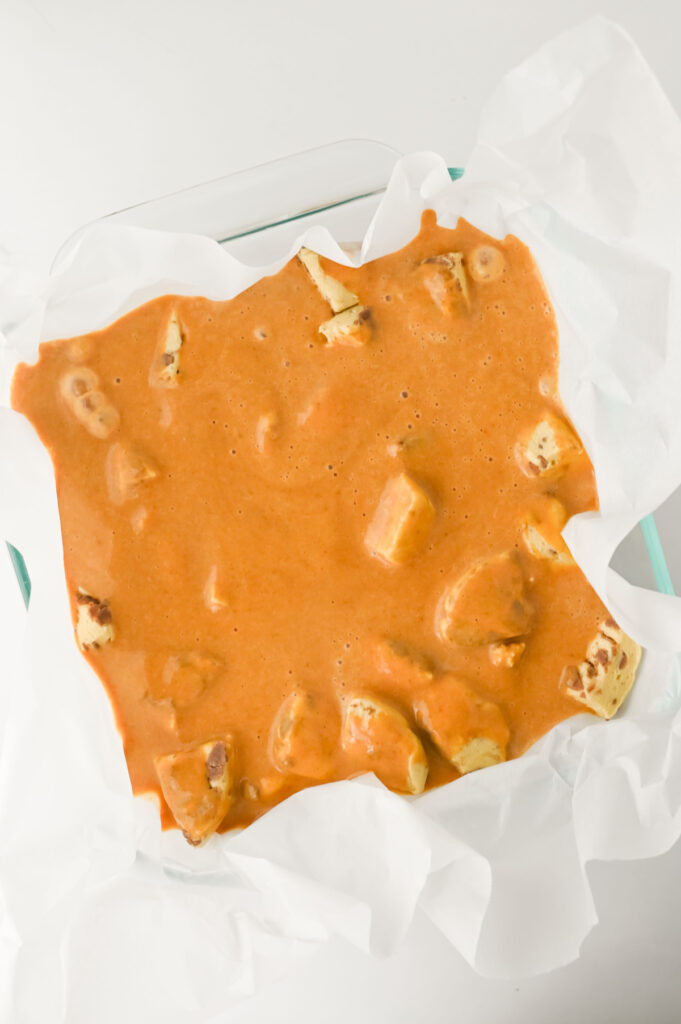 A glass dish lined with parchment paper contains cubes of paneer covered in an orange creamy sauce.