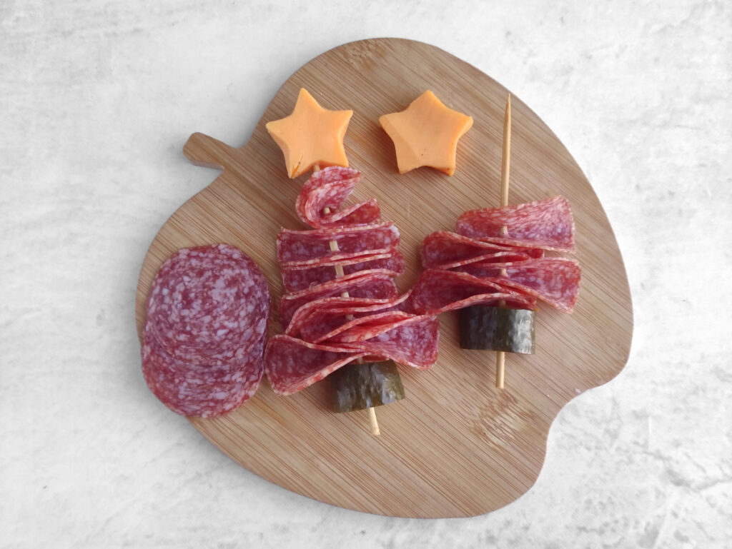 Two skewers with folded salami, cucumber slices, and cheddar cheese stars on a wooden apple-shaped board, with extra slices of salami on the side.