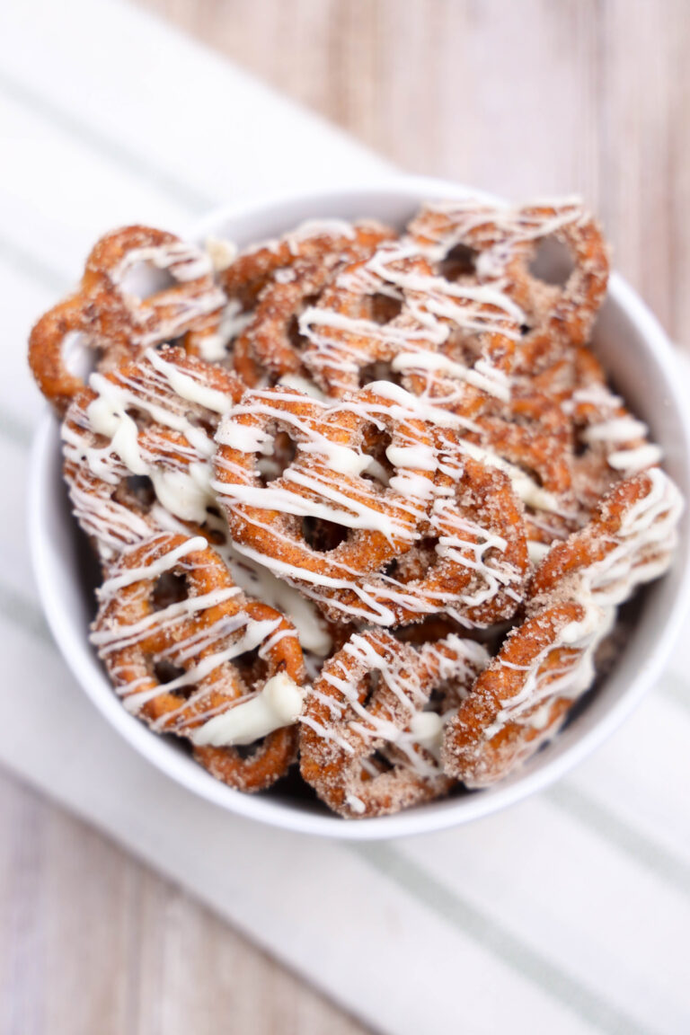 A white bowl filled with cinnamon roll pretzels coated in cinnamon sugar and drizzled with white chocolate, placed on a striped cloth.