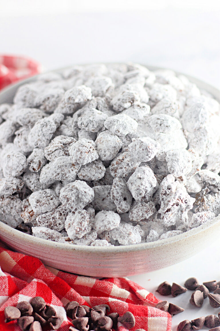 A bowl filled with powdered sugar-coated snack mix, also known as "puppy chow," sits on a red plaid cloth with chocolate chips scattered nearby.