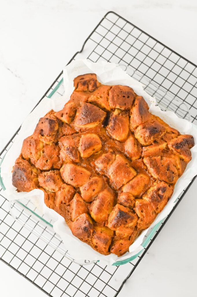 A baked monkey bread loaf with a golden-brown crust rests on parchment paper in a glass dish, placed on a black cooling rack.