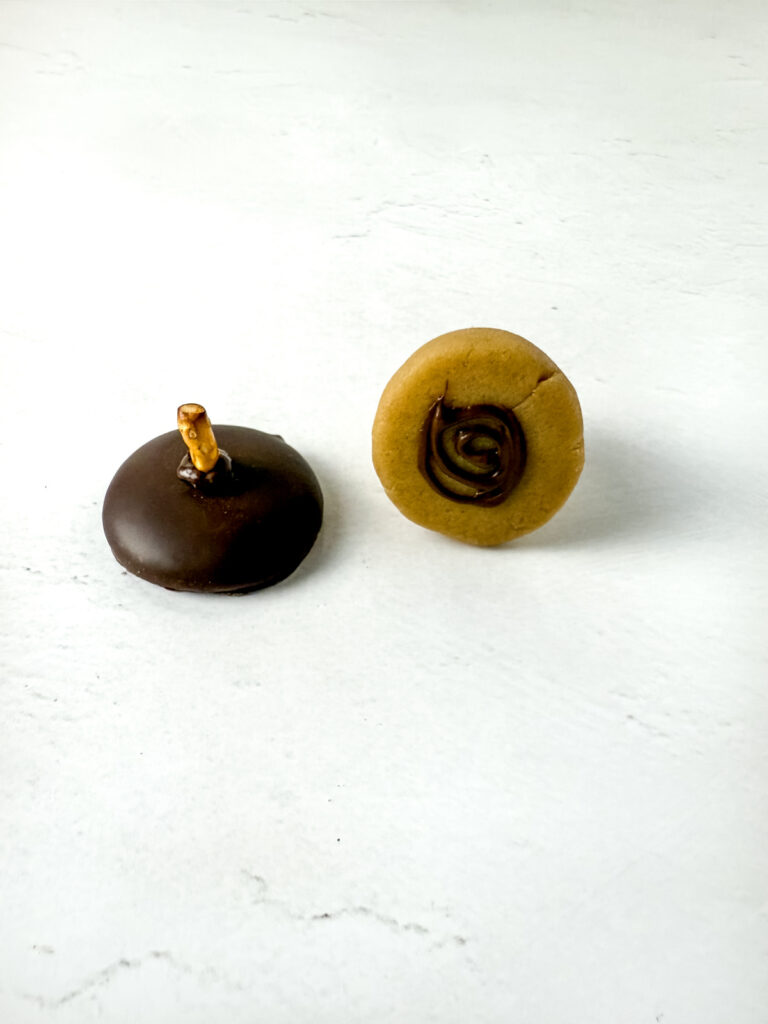 Two round cookies on a white surface; the left cookie is coated in chocolate with a pretzel stick, the right cookie is plain with a swirl of chocolate on top.