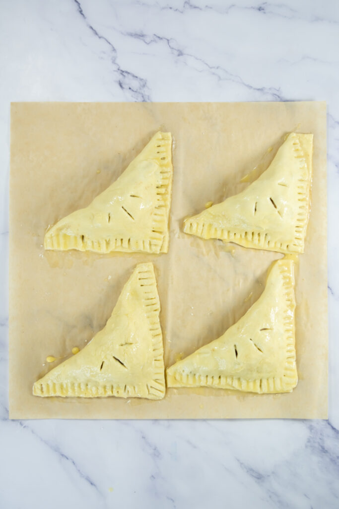 Four unbaked, triangular pastry turnovers with fork-sealed edges and small slits on top are arranged on a sheet of parchment paper on a marble surface.