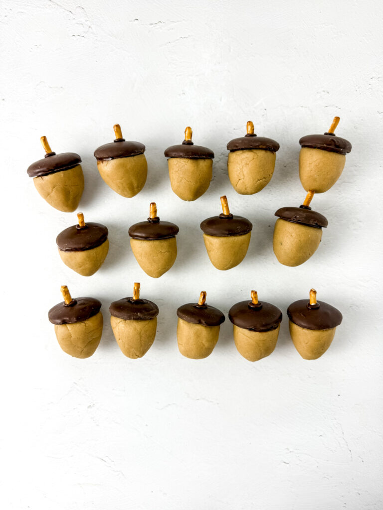 Fourteen acorn-shaped treats with brown tops and pretzel stick stems are arranged in rows on a white background.