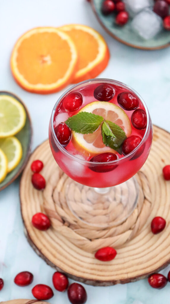 A glass of red beverage with ice, cranberries, lemon and orange slices, and a mint sprig, surrounded by loose cranberries and citrus slices on a wooden surface.