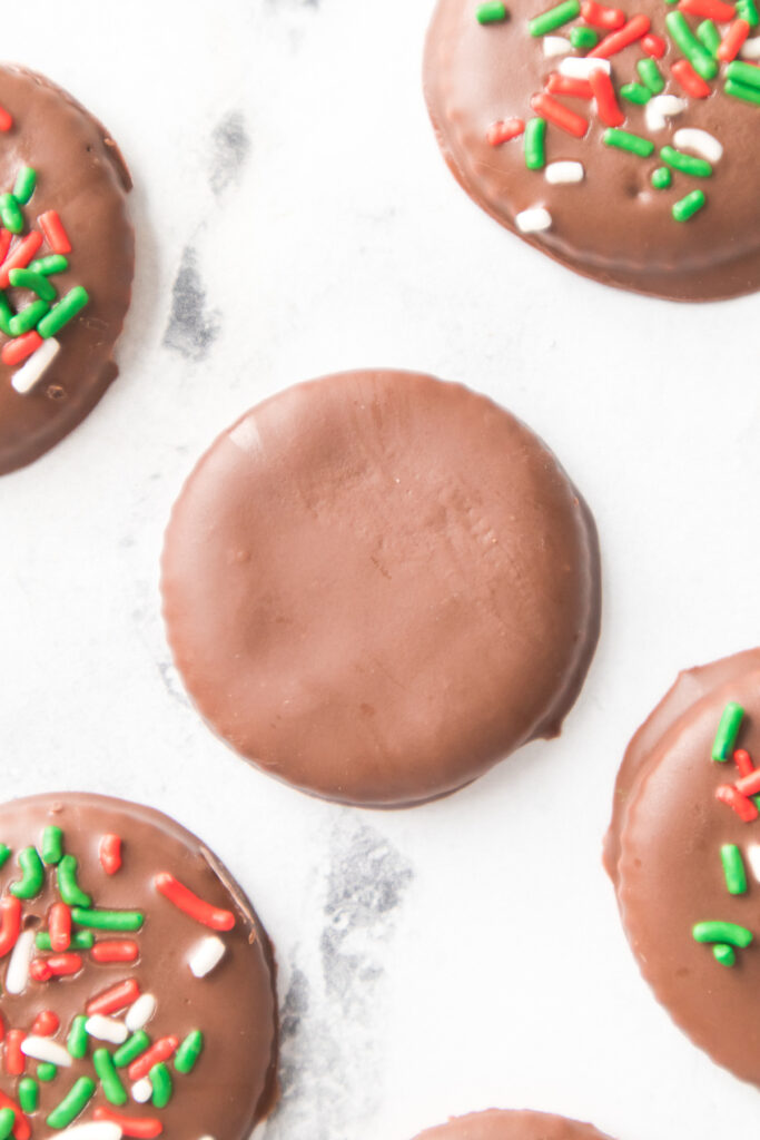 Chocolate-covered round cookies are arranged on a white surface, with some topped with red, green, and white sprinkles and one plain cookie in the center.
