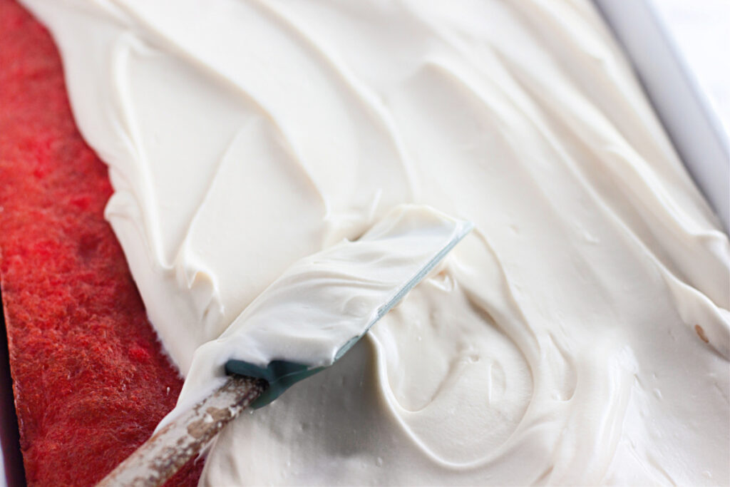 A spatula spreads a thick layer of white frosting over a red cake base in a rectangular pan.