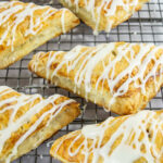 Six golden-brown, triangular pastries with white icing drizzle rest on a metal cooling rack.