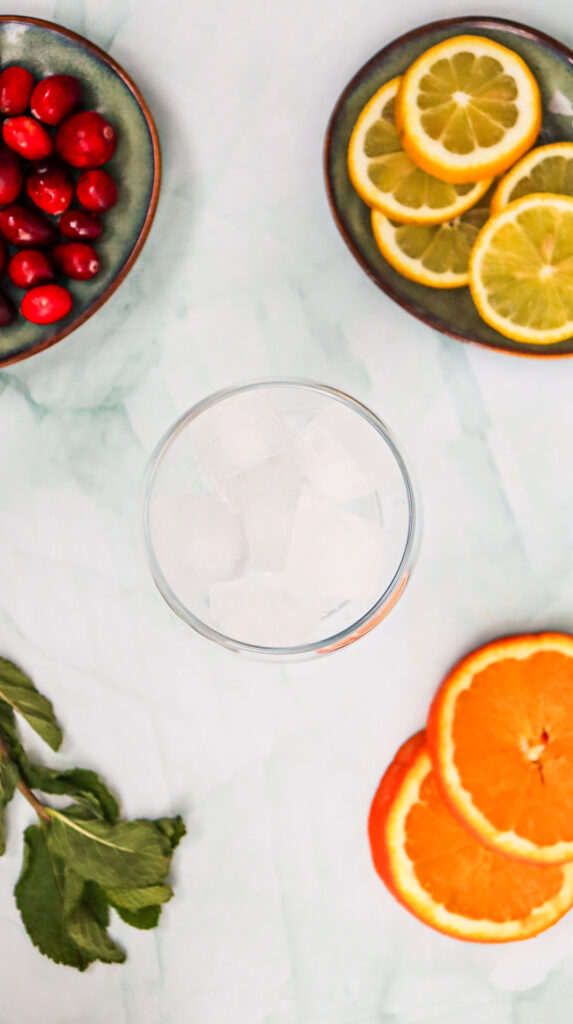 A glass with ice cubes surrounded by plates of cranberries, lemon and lime slices, orange slices, and fresh mint on a light surface.