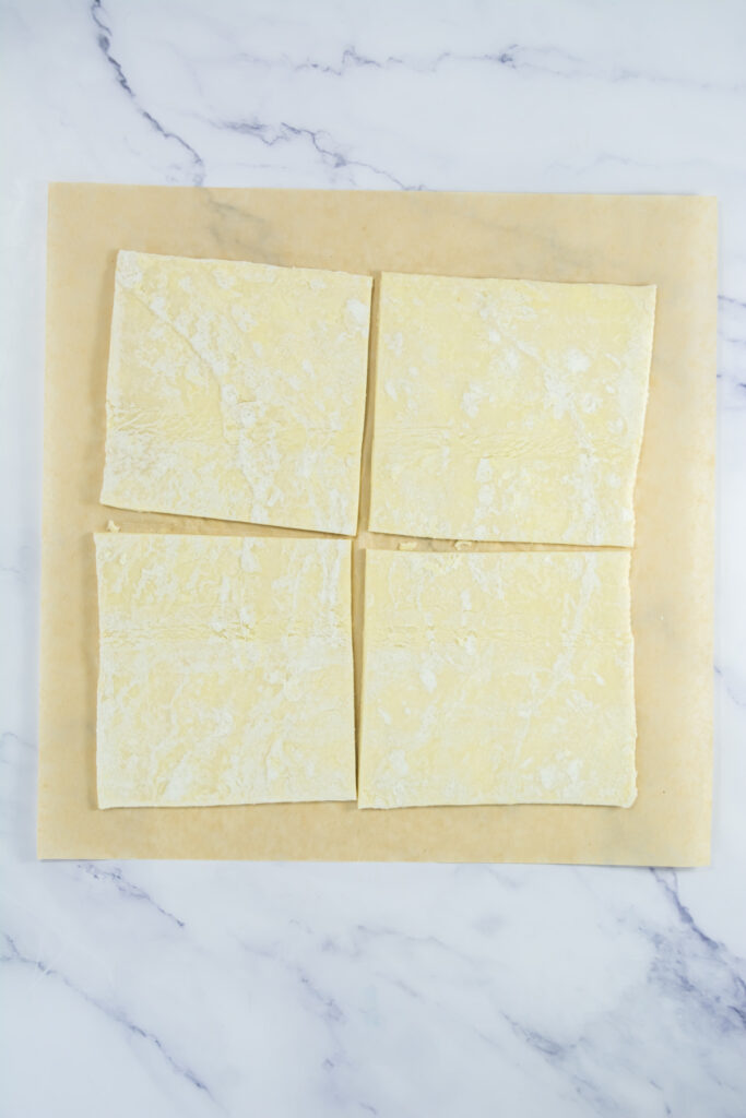 Four uncooked puff pastry squares are arranged on a sheet of parchment paper atop a marble surface.