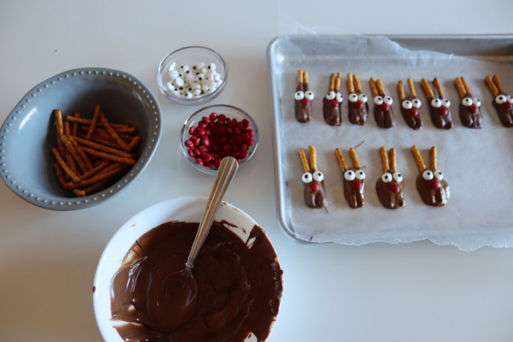 Chocolate-covered cookies with candy eyes, red nose candies, and pretzel antlers arranged on a baking tray. Ingredients are in bowls nearby on a white surface.