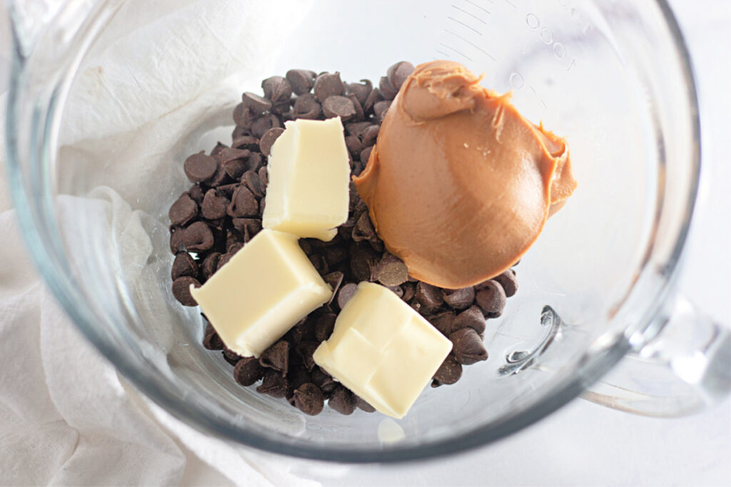 A glass bowl containing chocolate chips, three cubes of butter, and a scoop of peanut butter, viewed from above.
