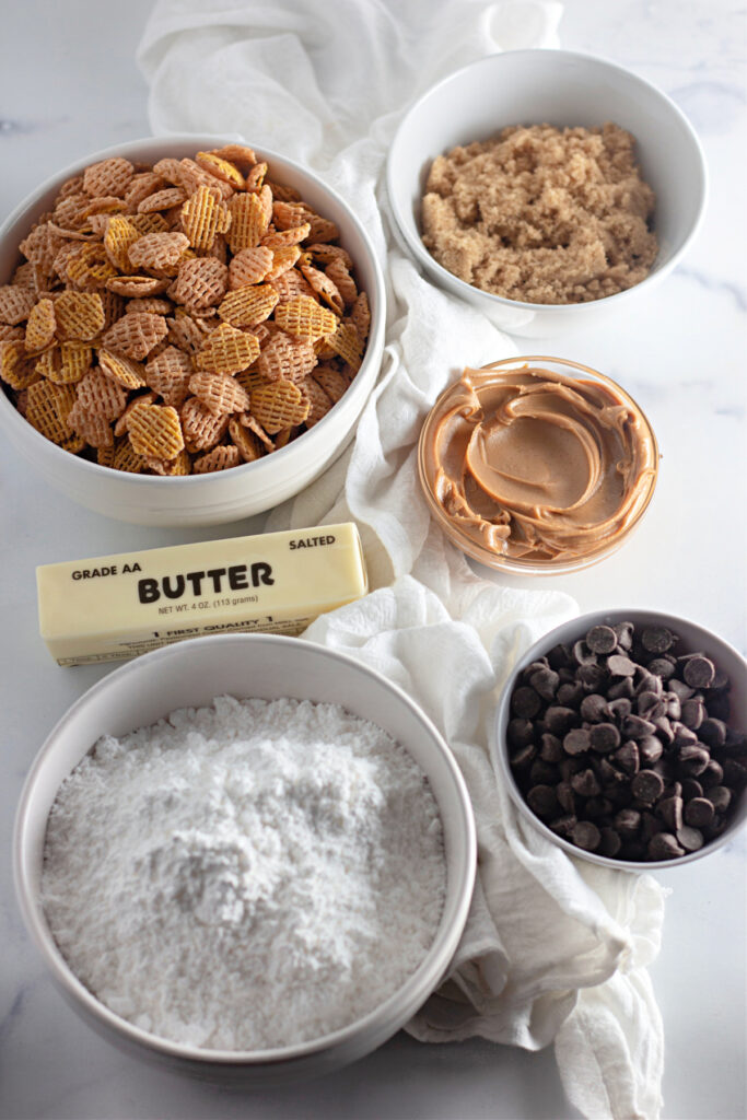 Bowls of Chex cereal, brown sugar, powdered sugar, peanut butter, chocolate chips, and a stick of salted butter arranged on a white surface.