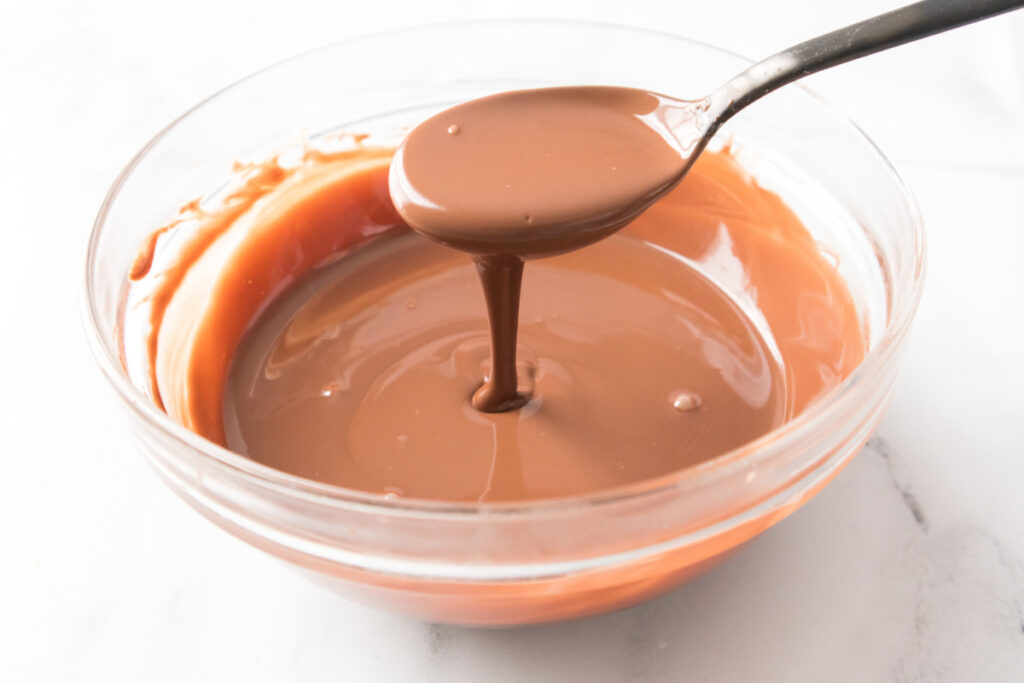 A spoon holds up and drizzles melted milk chocolate over a clear glass bowl filled with more melted chocolate.