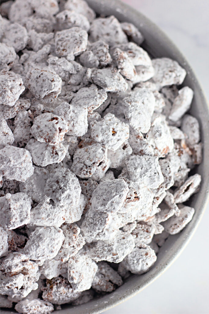 A close-up of a bowl filled with powdered sugar-coated snack clusters, commonly known as puppy chow or muddy buddies.