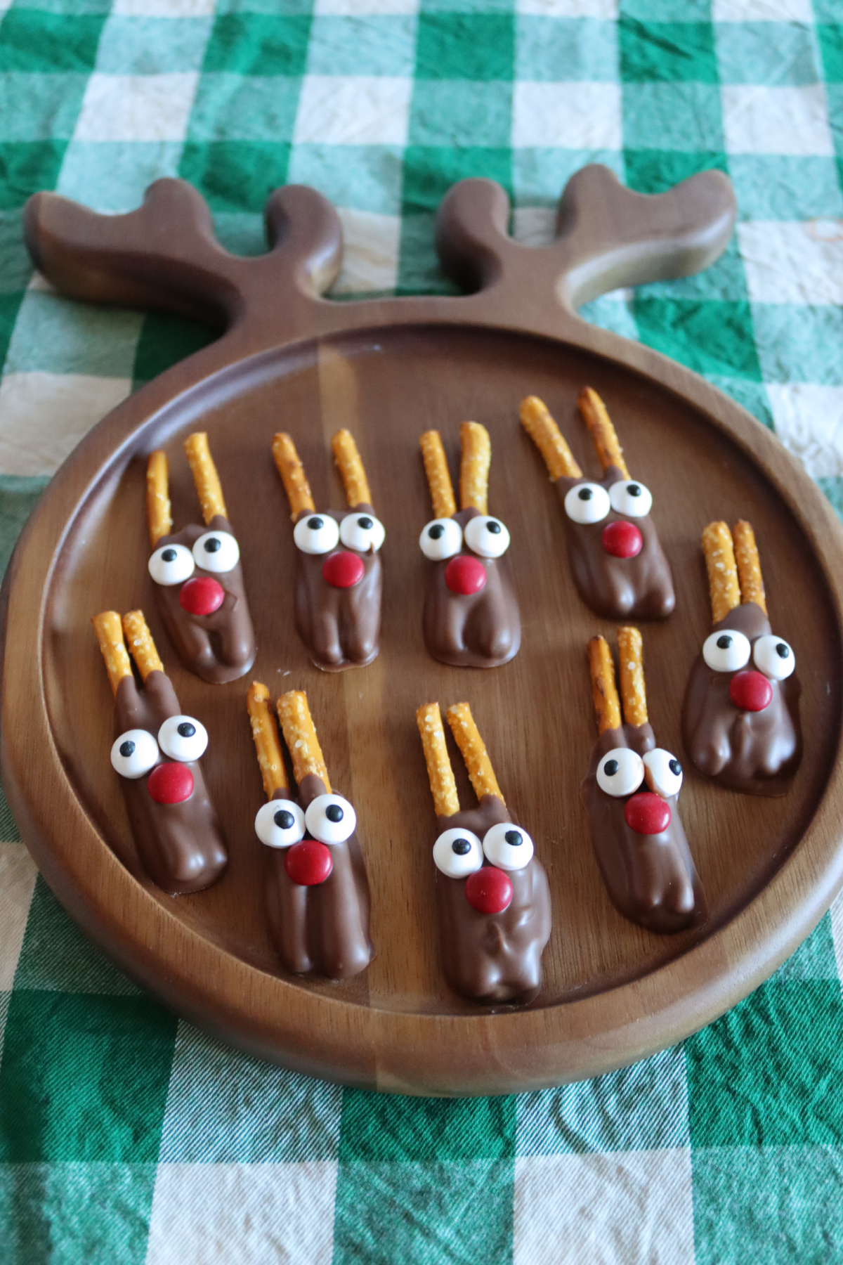 A round wooden tray with eight pretzel reindeers—chocolate-covered treats decorated with candy eyes, red noses, and pretzel sticks—sits atop a green and white checkered tablecloth.