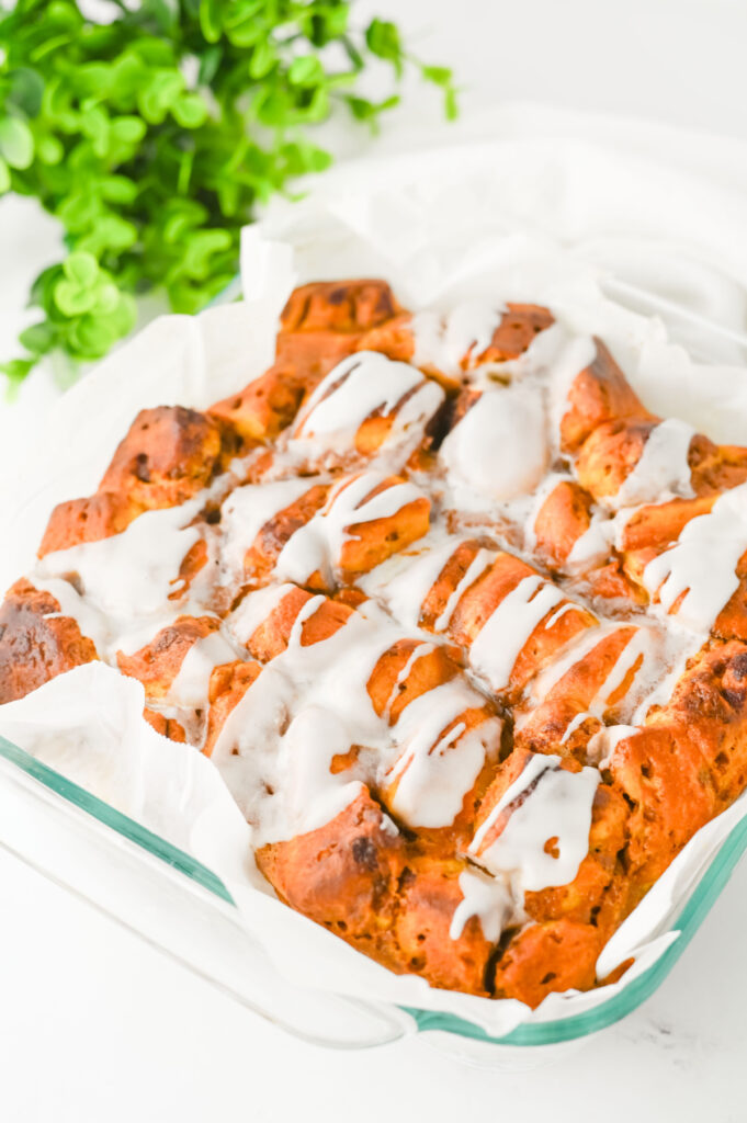 A glass baking dish with iced cinnamon roll bites on parchment paper, with a green plant in the background.