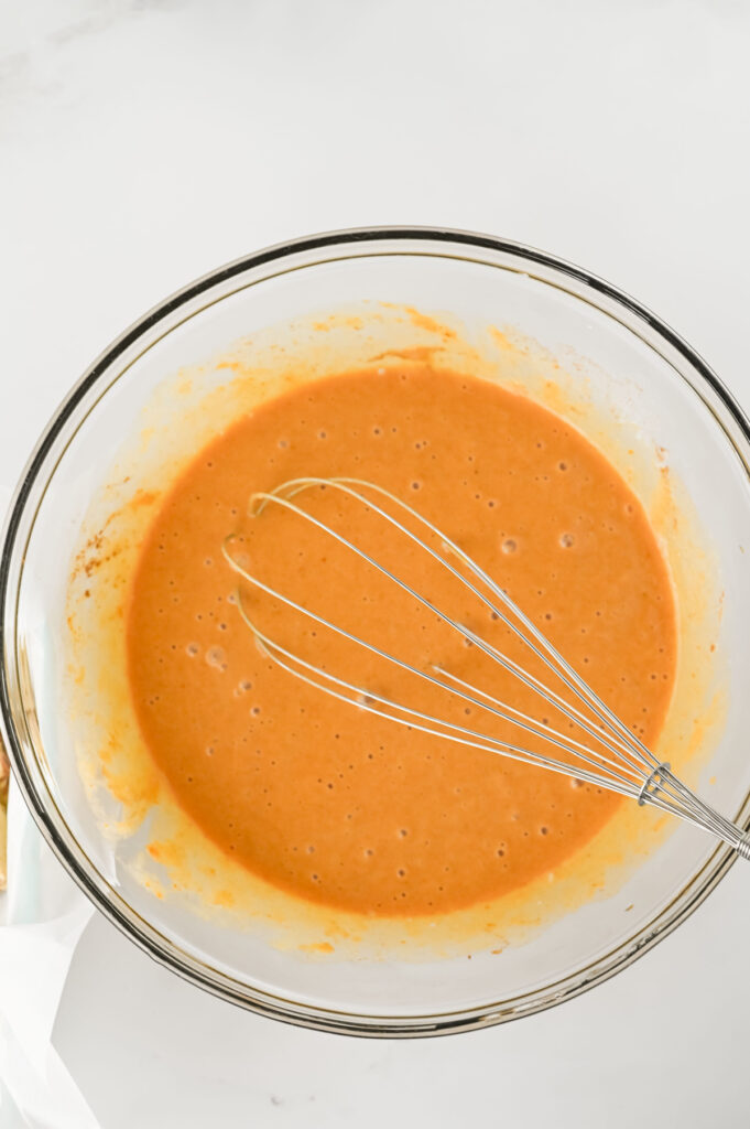 A metal whisk rests in a glass bowl filled with orange batter on a white surface.