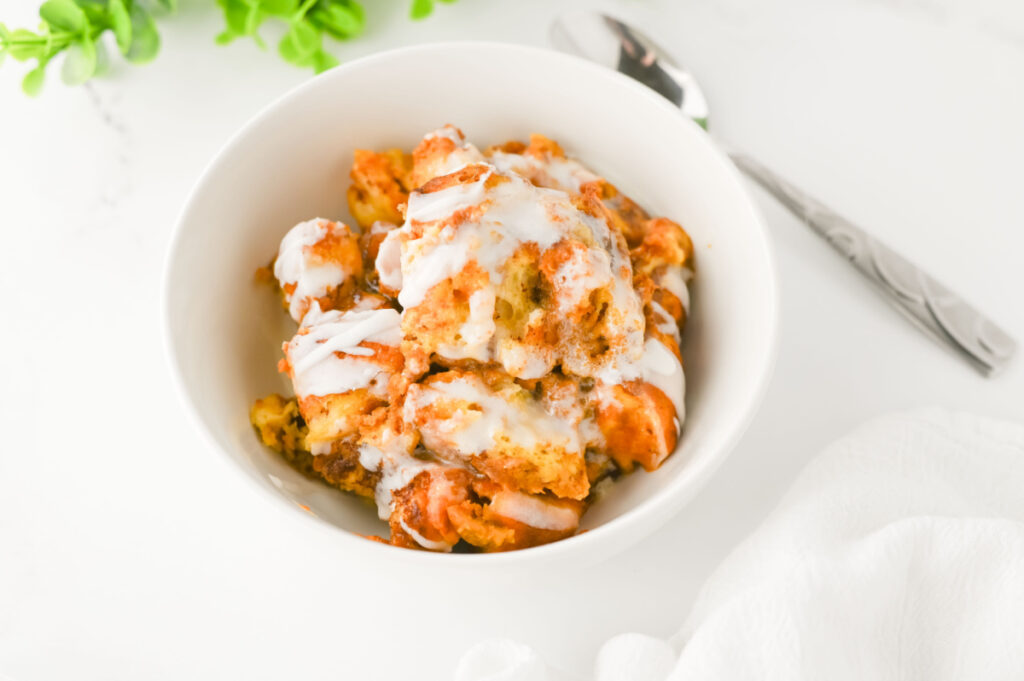 A white bowl filled with bread pudding drizzled with white icing, placed on a white surface next to a spoon and a white napkin.