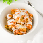 A white bowl filled with bread pudding drizzled with white icing, placed on a white surface next to a spoon and a white napkin.