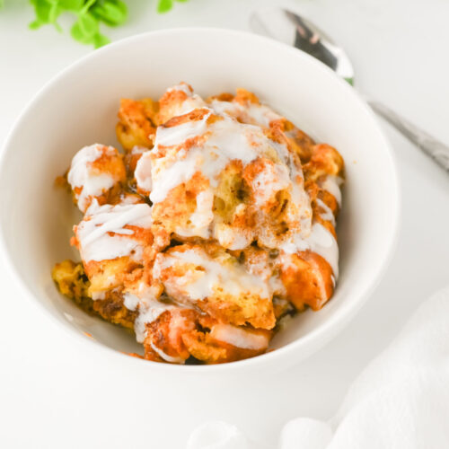 A white bowl filled with bread pudding drizzled with white icing, placed on a white surface next to a spoon and a white napkin.