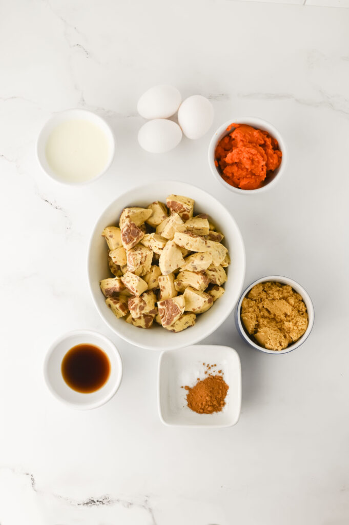 Bowls with cubed bread, brown sugar, milk, pumpkin puree, spices, vanilla extract, and three eggs are arranged on a white marble surface.