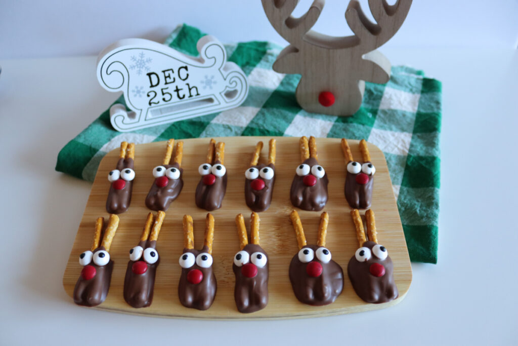 A wooden board with twelve chocolate-covered treats shaped like reindeer, made with pretzel antlers, candy eyes, and red noses, sits on a table with holiday decorations in the background.