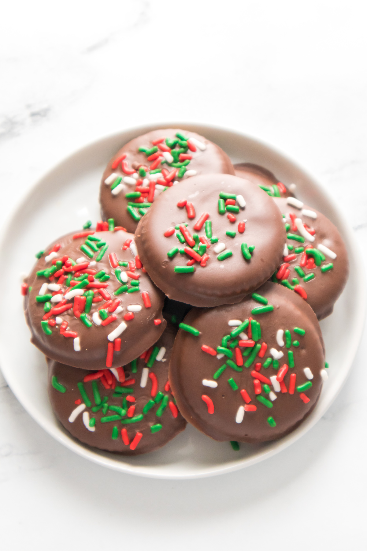A white plate with chocolate-covered cookies topped with red, green, and white sprinkles, placed on a white surface.