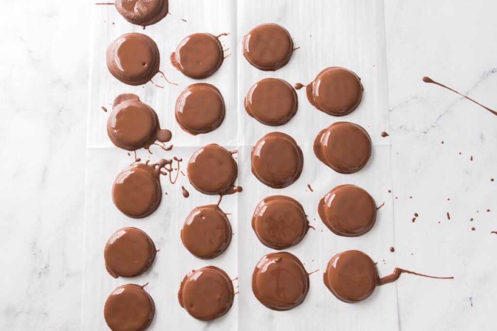 Rows of round cookies coated in chocolate are arranged on parchment paper, with some chocolate drips and smudges visible around them.